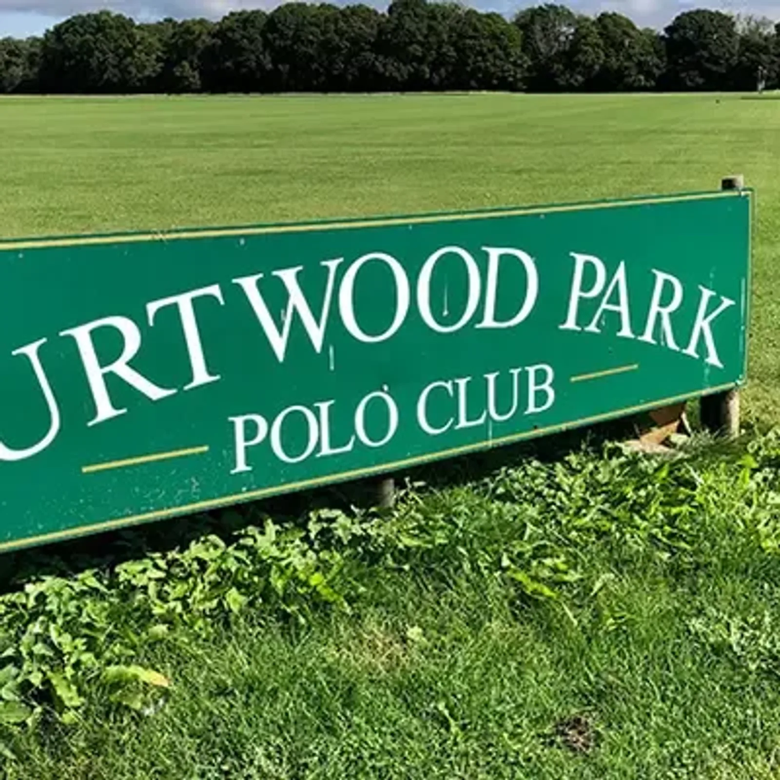 A green sign reads "Hurtwood Park Polo Club" in white letters, set against a lush green field with trees in the background.