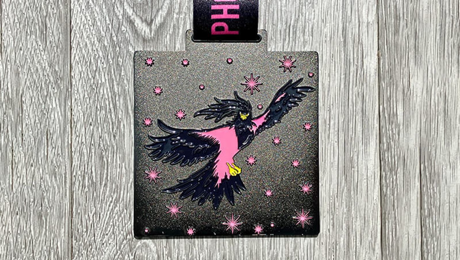 A square medal with a metallic finish depicts a stylized bird with black and pink feathers in mid-flight. The background is adorned with pink star-like shapes. The medal hangs from a black ribbon with the word "PHOENIX" in pink letters. The wood texture backdrop enhances the design.