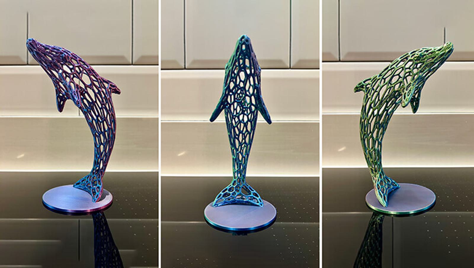 Three photos of a digital dolphin sculpture with a geometric, lattice design. Each appears in different colors: purple, blue, and green, mounted on a circular base, set against a neutral background.