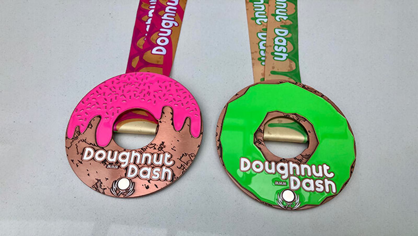 Two doughnut-shaped medals with pink and green icing designs. Each medal reads "Doughnut Dash" and features colorful ribbons. The pink medal is on the left, and the green medal is on the right, both resting on a white surface.