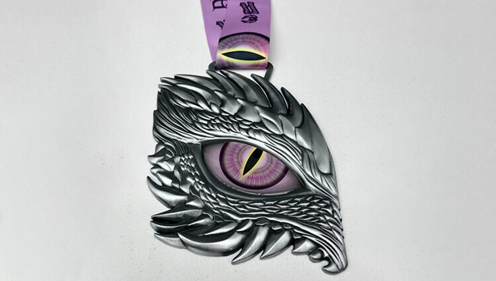 A metallic medal shaped like a dragon's head with intricate scale details and a prominent purple, reptilian eye at the center. The medal is attached to a purple ribbon.
