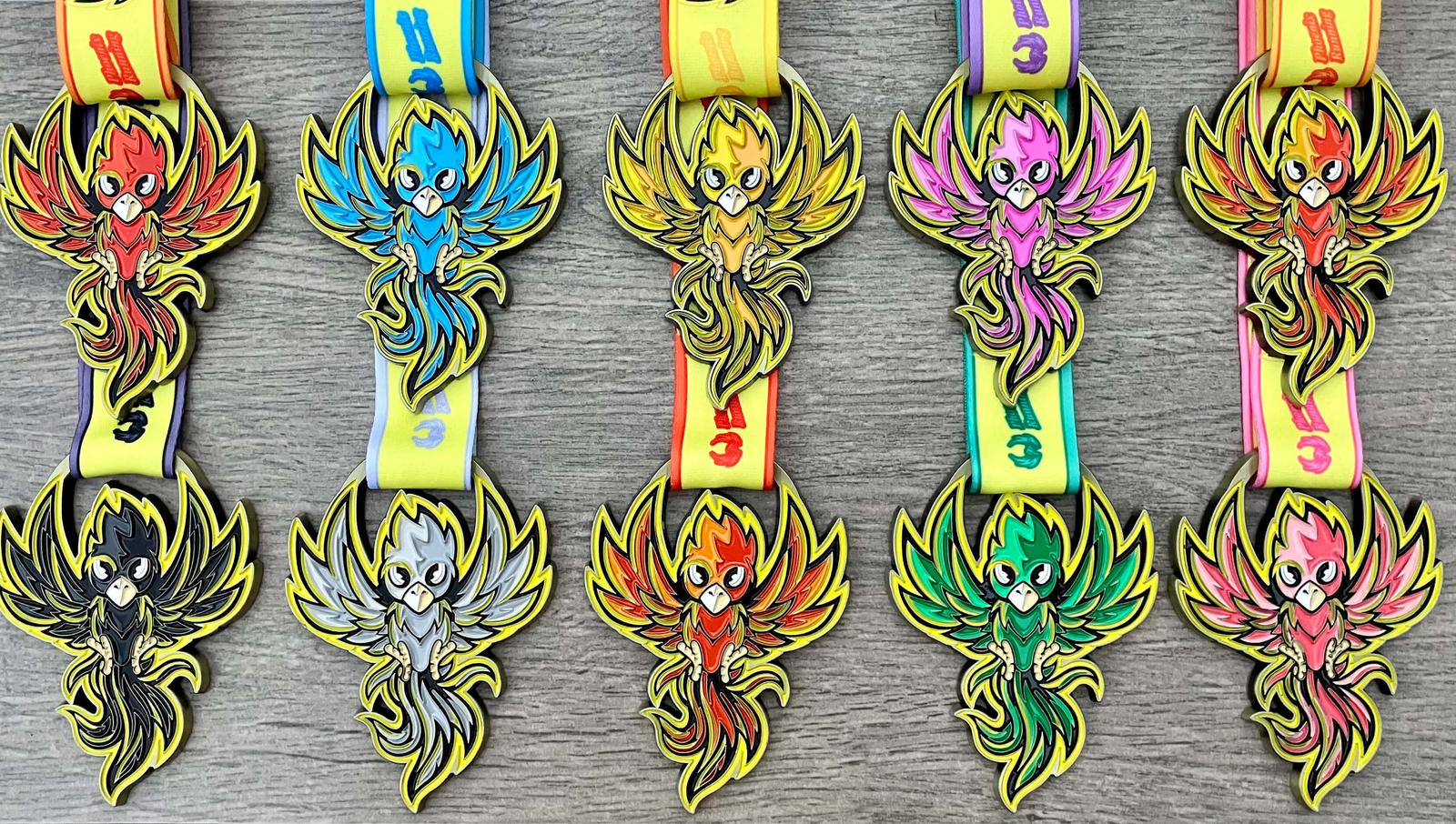 A row of five colorful medals featuring phoenix designs in red, blue, yellow, pink, and green, each with a matching ribbon. The medals are arranged on a wooden surface.