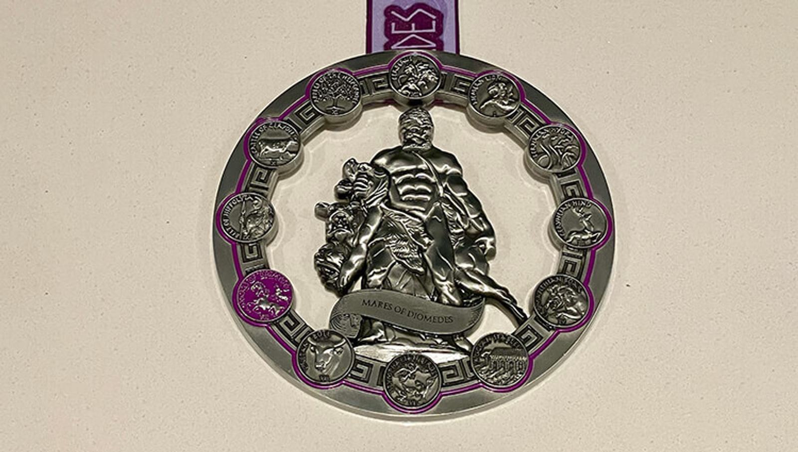A silver marathon medal features a muscular figure with an animal, surrounded by twelve smaller engraved designs and a purple ribbon. The central figure stands on a base reading "MARCUS OCTAVIUS" against a beige background.