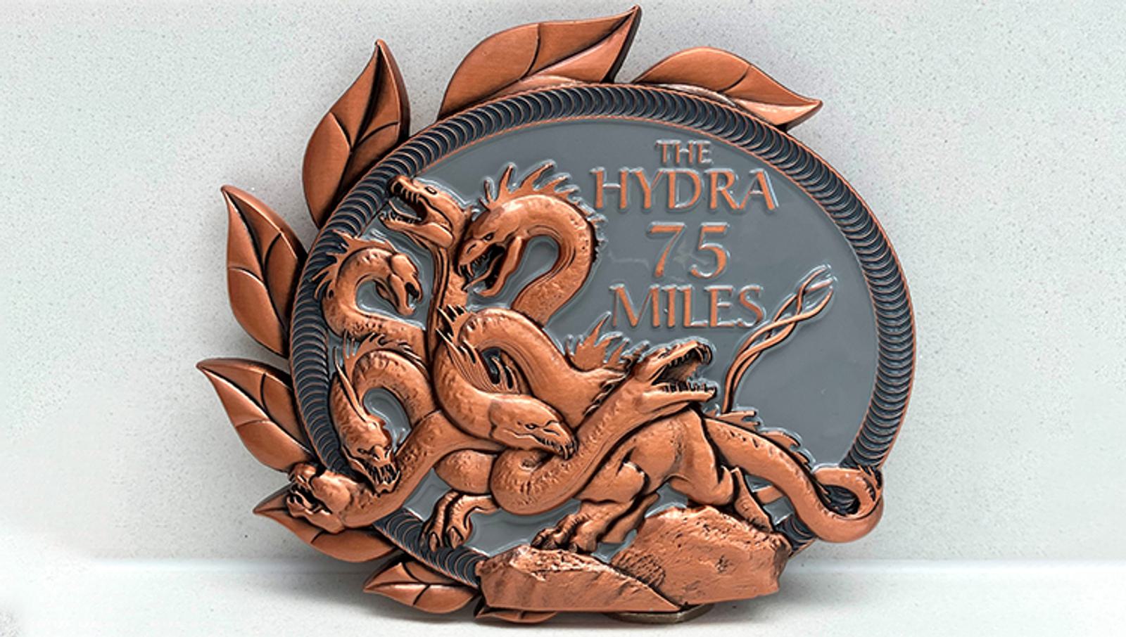 A decorative medal with a bronze hydra design encircling the words "The Hydra 75 Miles." The hydra has multiple heads and is surrounded by leaf motifs, set against a textured background.