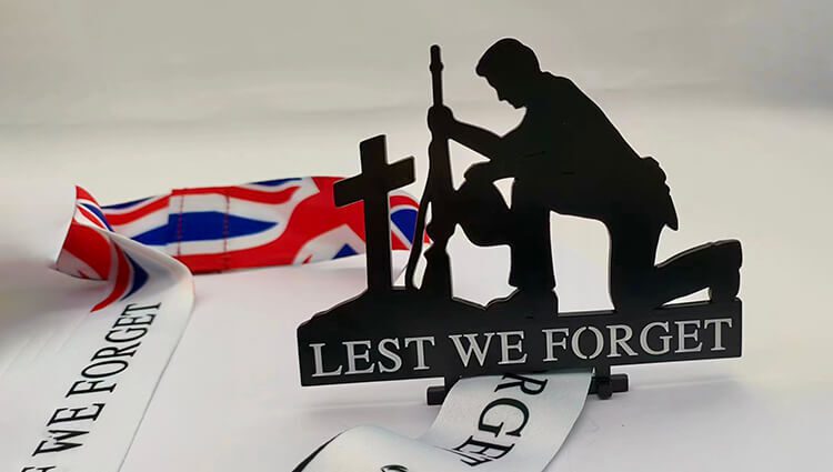 Silhouette of a soldier kneeling beside a cross with a rifle, set against a Union Jack ribbon. The words "LEST WE FORGET" are displayed prominently in front.