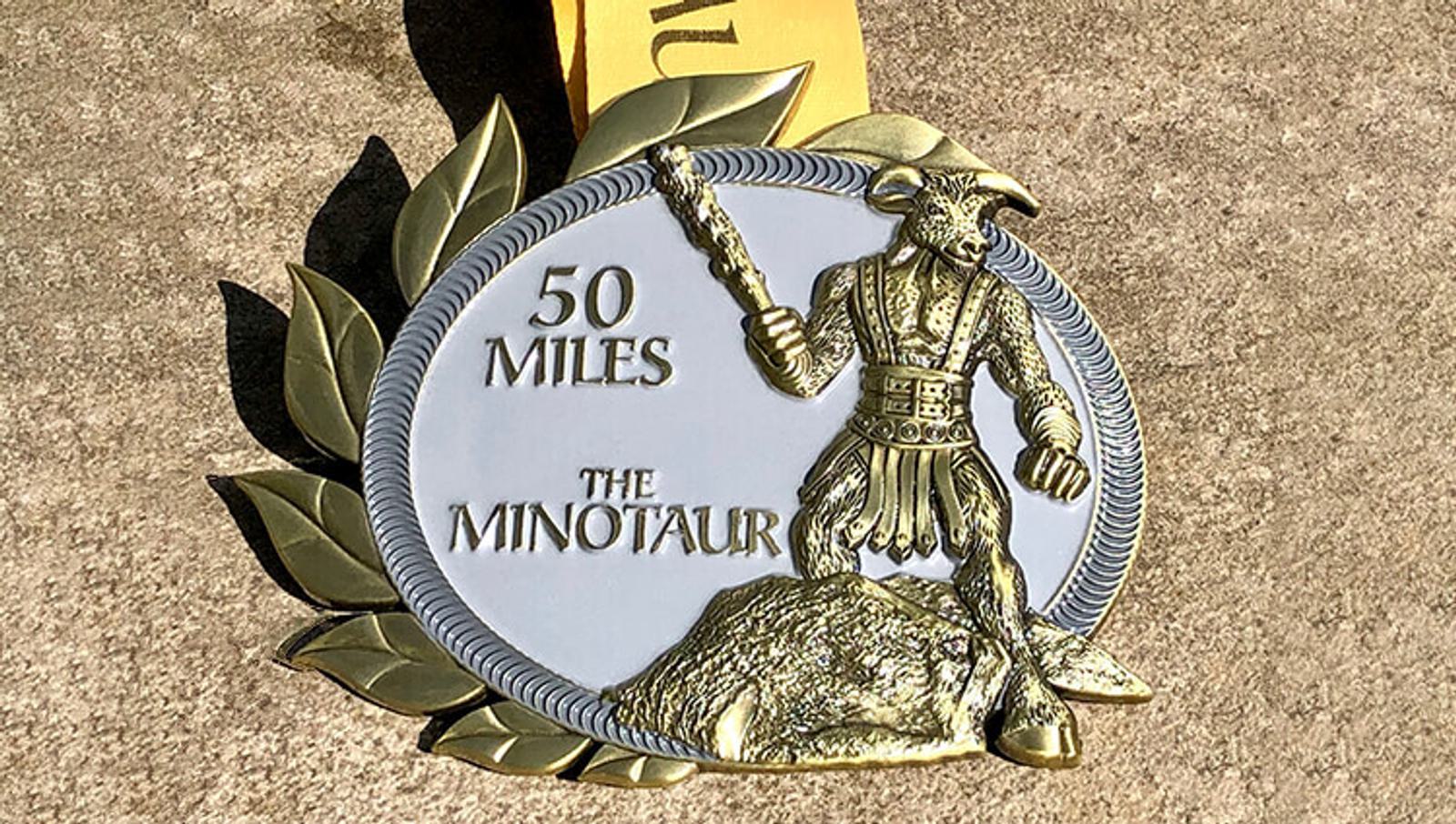 An oval-shaped medal featuring a detailed Minotaur holding a club, standing on a rock. The text "50 Miles" and "The Minotaur" is engraved, with a leaf pattern on the left. The medal is attached to a yellow ribbon.