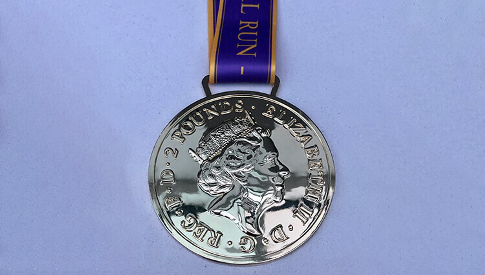 A gold-colored medal resembling a coin with an embossed profile of Queen Elizabeth II. The medal is attached to a purple ribbon with gold lettering partially visible, possibly denoting an event or run. The background is a plain light surface.