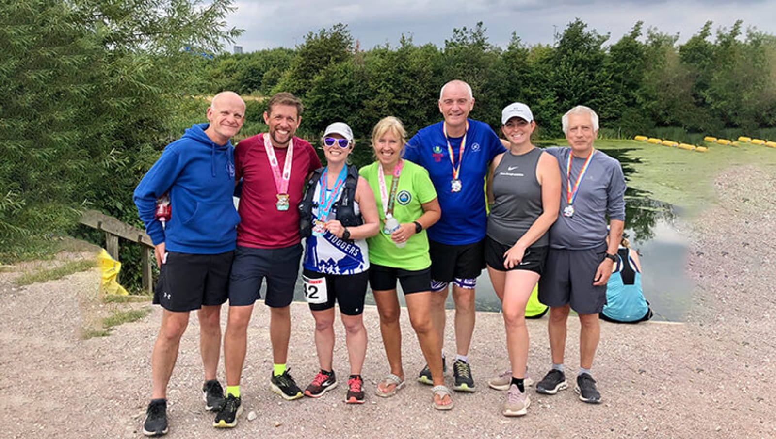 Seven people wearing athletic clothes and medals stand smiling together outdoors near a pond, with trees and greenery in the background. Some hold race bibs, suggesting they've completed a race or sporting event.