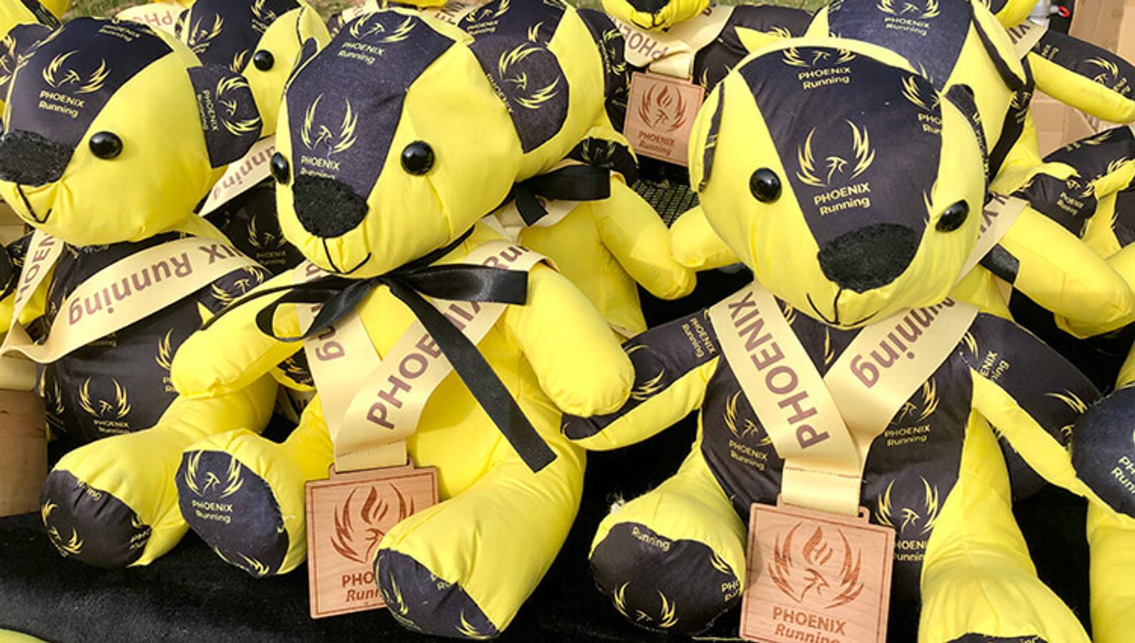 A group of yellow and black teddy bears, each wearing a Phoenix Running sash and a wooden Phoenix Running medal, are arranged closely together. The bears have black patches on their ears, paws, and around their eyes.