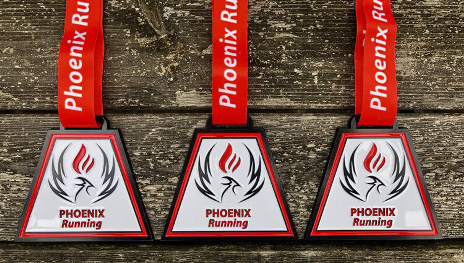 Three triangular medals with red and black designs hang on a wooden background. Each medal features a stylized flame and bird logo with the text "PHOENIX Running." The ribbons are red with "Phoenix Running" printed in white.