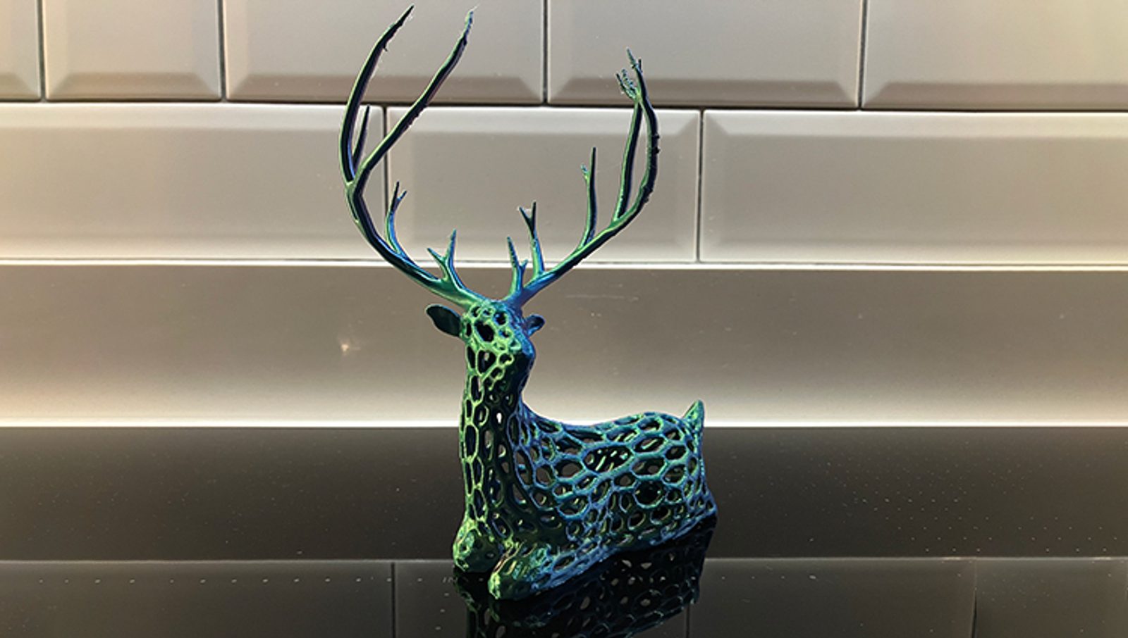 A decorative sculpture of a stag with large antlers, featuring a blue and green lattice pattern, sits on a shiny black surface in front of white tiled walls.