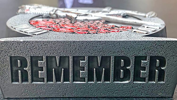 A textured gray stone plaque with the word "REMEMBER" engraved on it. Above, there is a circular design with a layer of red stones and silver metallic elements arranged decoratively.