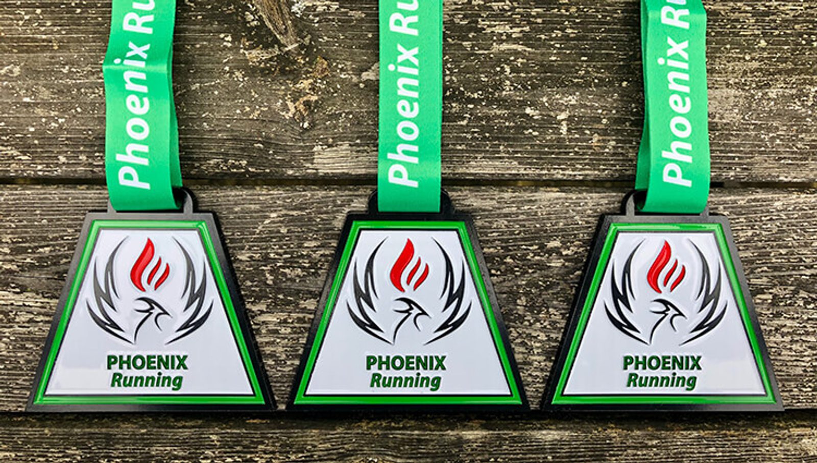 Three triangular medals with green lanyards displaying the text "Phoenix Running". Each medal features a flame design with a phoenix outline, the word "PHOENIX" in bold green, and "Running" below it. They are placed on a rustic wooden surface.