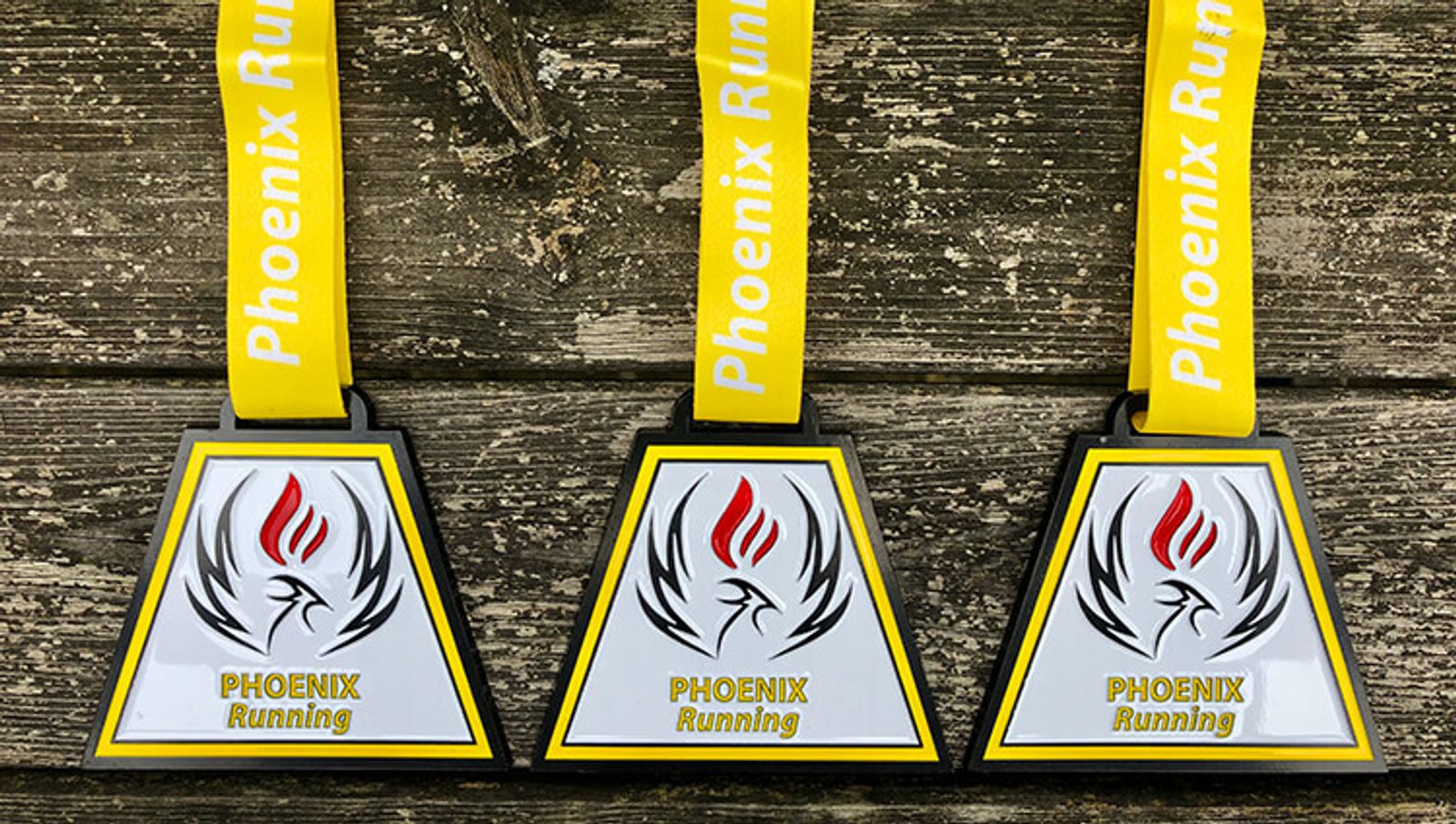 Three triangular medals with a yellow and white ribbon reading "Phoenix Running" are displayed on a wooden surface. The front of each medal features a stylized phoenix in black outline with red and white accents, and the text "PHOENIX Running.