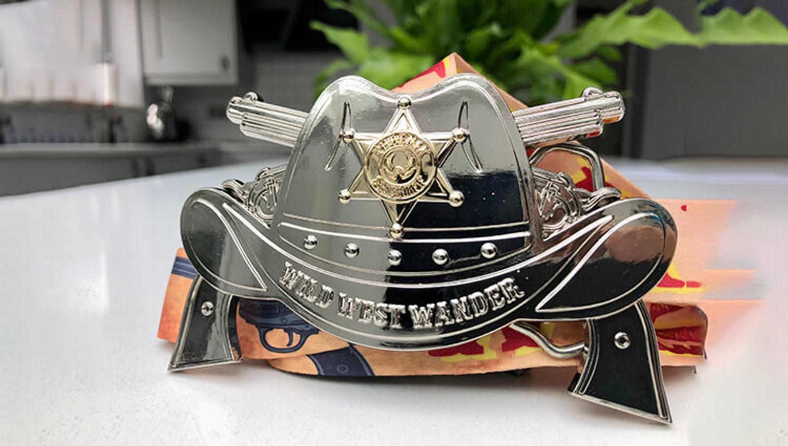 A shiny cowboy hat-shaped belt buckle with intricate detailing and a sheriff's star in the center, featuring the words "WILD WEST WANDER." It is displayed on a kitchen countertop with blurred greenery in the background.