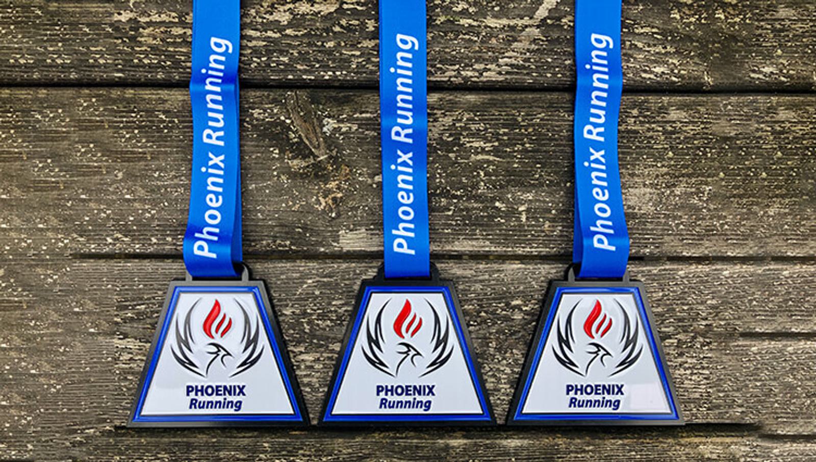 Three triangular medals with blue ribbons displaying "Phoenix Running" are laid out on a wooden surface. Each medal features a phoenix graphic above the text.