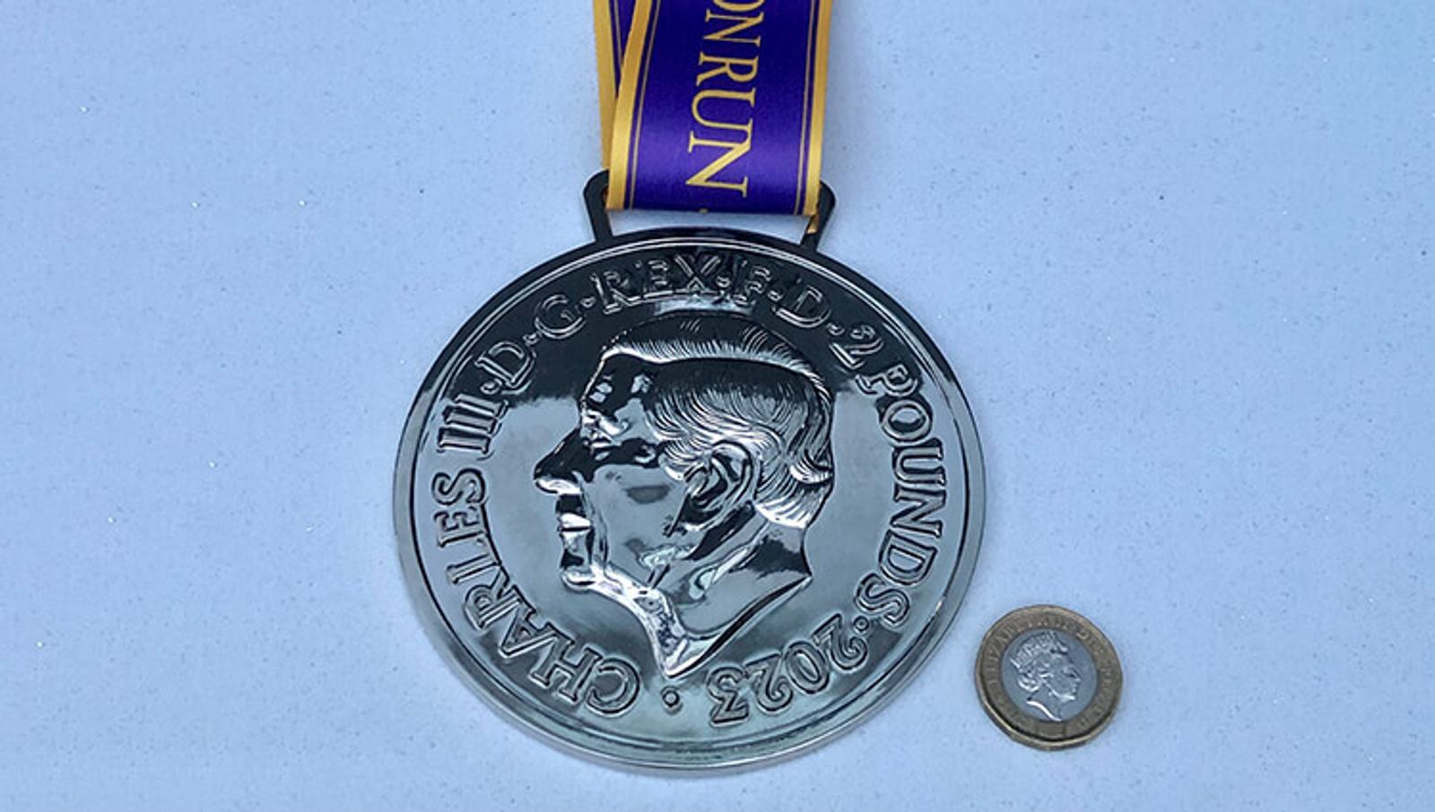 A large silver medal featuring the profile of King Charles III with inscriptions "CHARLES III, D G, REX, F D, 2 POUNDS, 2022" sits on a white surface. The medal is attached to a purple and yellow ribbon with the word "MONRUN." A small coin is placed beside it for scale.