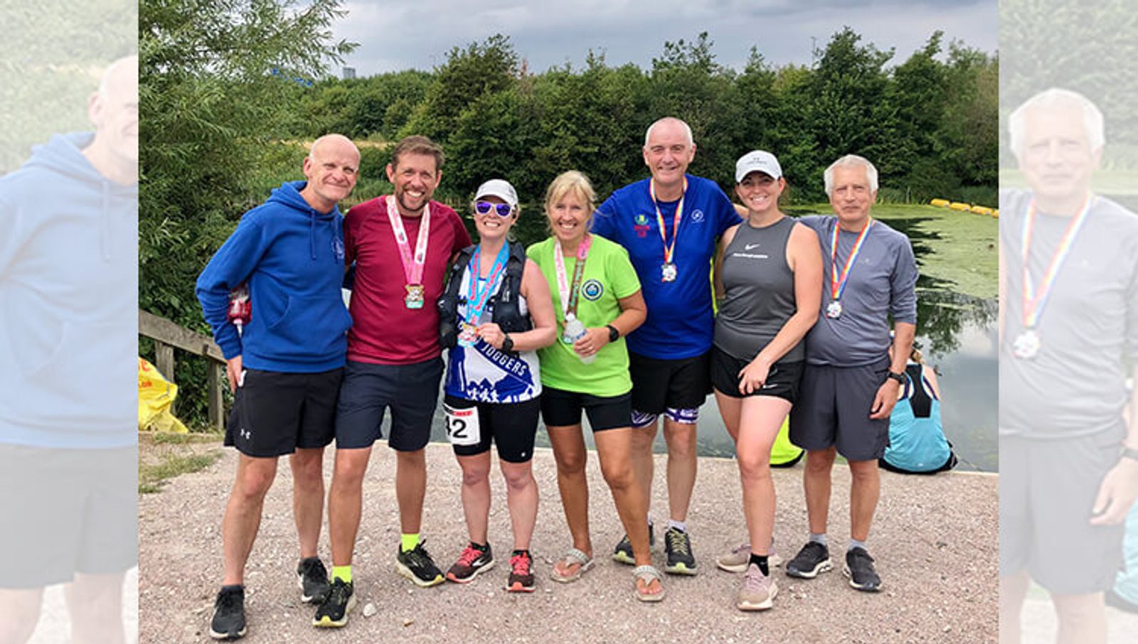 A group of seven people, wearing athletic clothing and race medals, stand side by side outdoors on a gravel path near a body of water. They are smiling and posing for the camera with trees and greenery in the background.