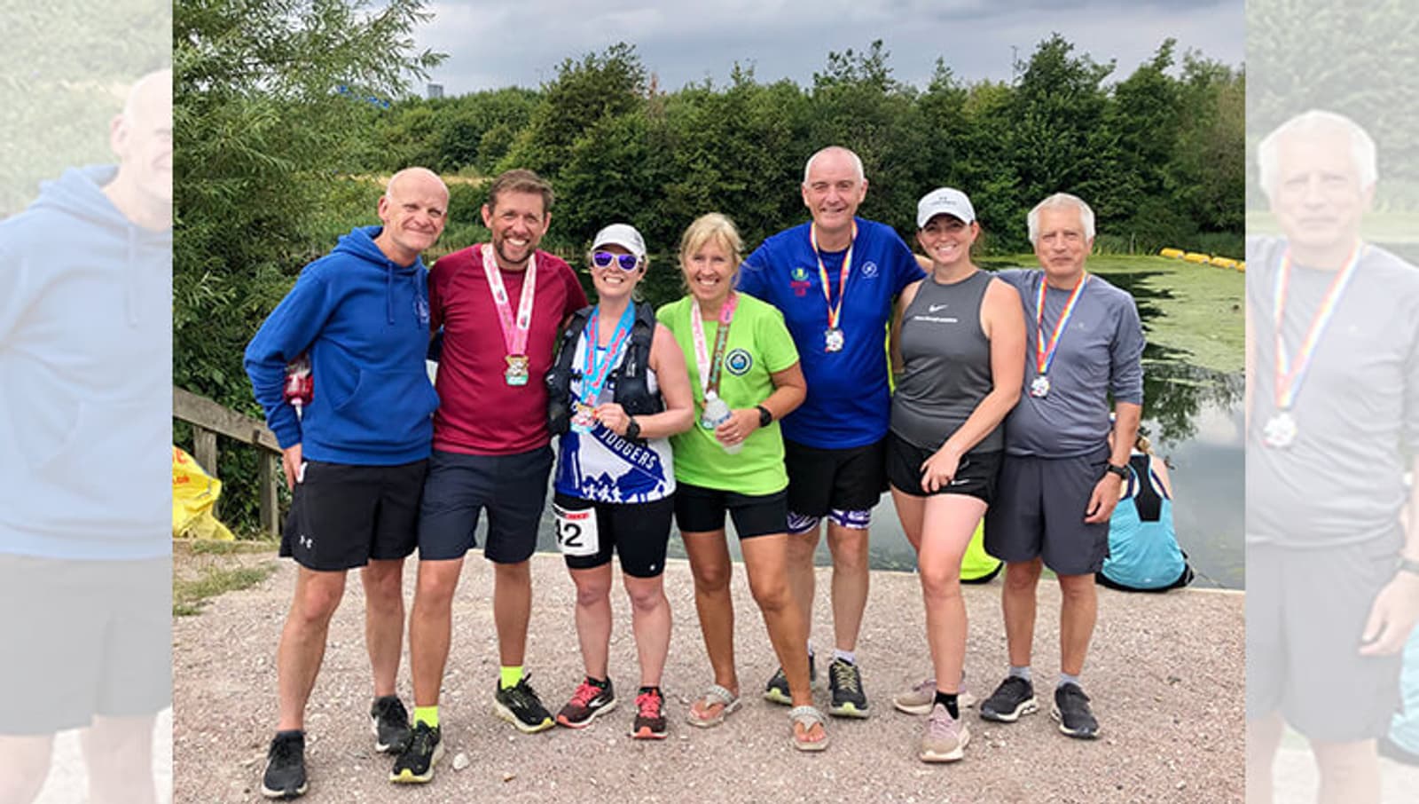 A group of seven people are standing outdoors, smiling at the camera. They are dressed in athletic gear and have medals around their necks, suggesting they participated in an athletic event. In the background, there is greenery and a body of water.