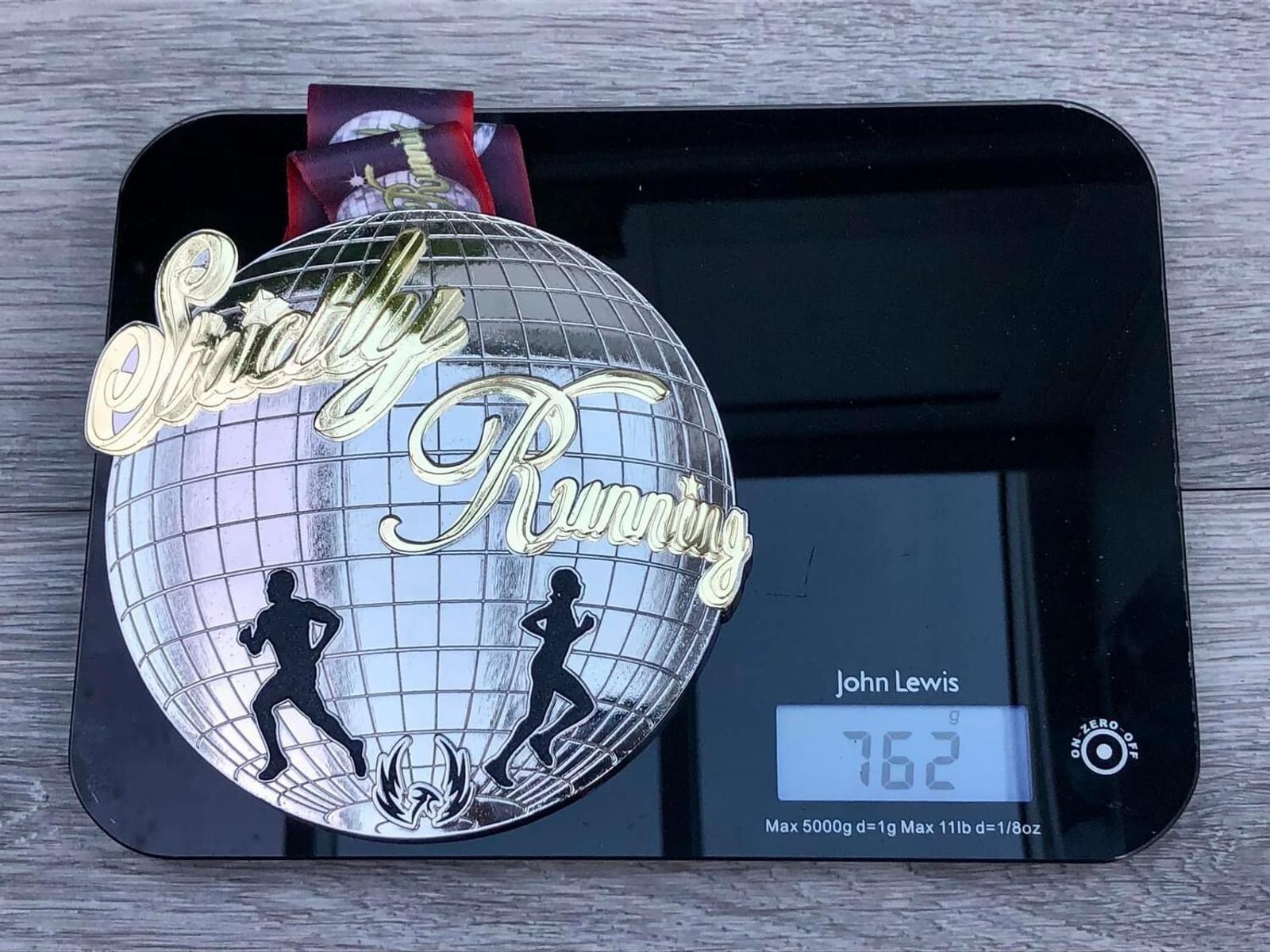 A "Strictly Running" medal with silhouettes of a runner and dancer is placed on a digital scale, showing a weight of 76.2 grams. The medal features a mirror ball design, and the scale has the brand name John Lewis on its surface.