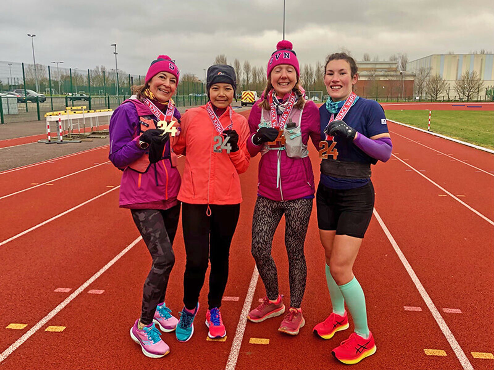 Four women, dressed in colorful athletic gear, stand together on an outdoor running track, smiling and holding medals. They appear to be celebrating their achievement in a race or running event. The weather is overcast, and they are wearing hats and gloves.