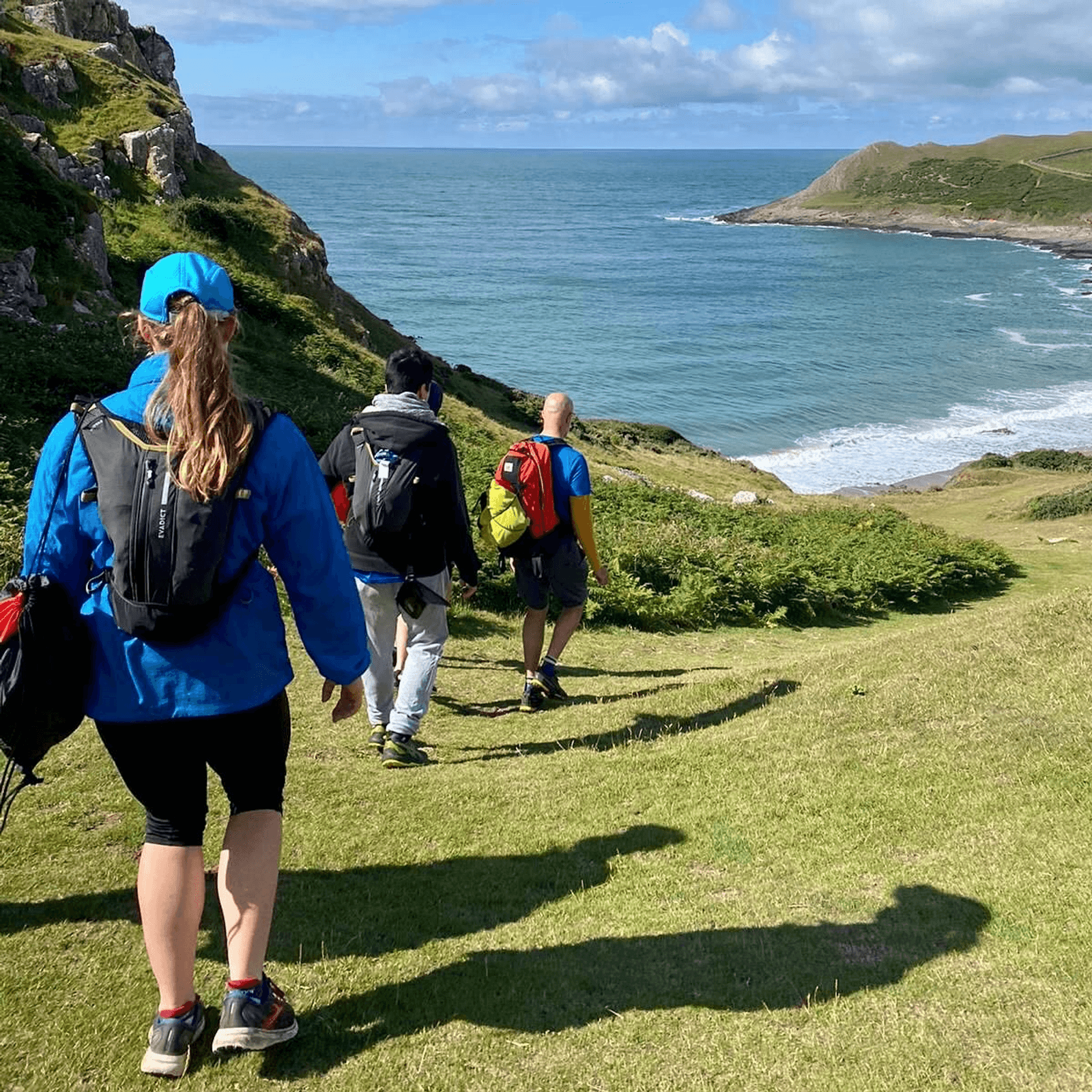 A group of people hiking down a grassy path towards a coastline. The sea is visible in the background under a partly cloudy sky. The hikers are dressed in outdoor gear, including backpacks and hats. The landscape features cliffs and greenery.