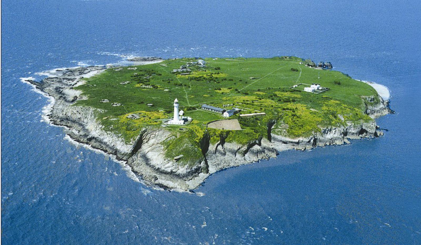 Aerial view of a grassy island surrounded by blue ocean waters. The island features a white lighthouse near the shore, several buildings, and scattered trees. Steep rocky cliffs define the island's perimeter, and a small sandy beach is visible in the background.