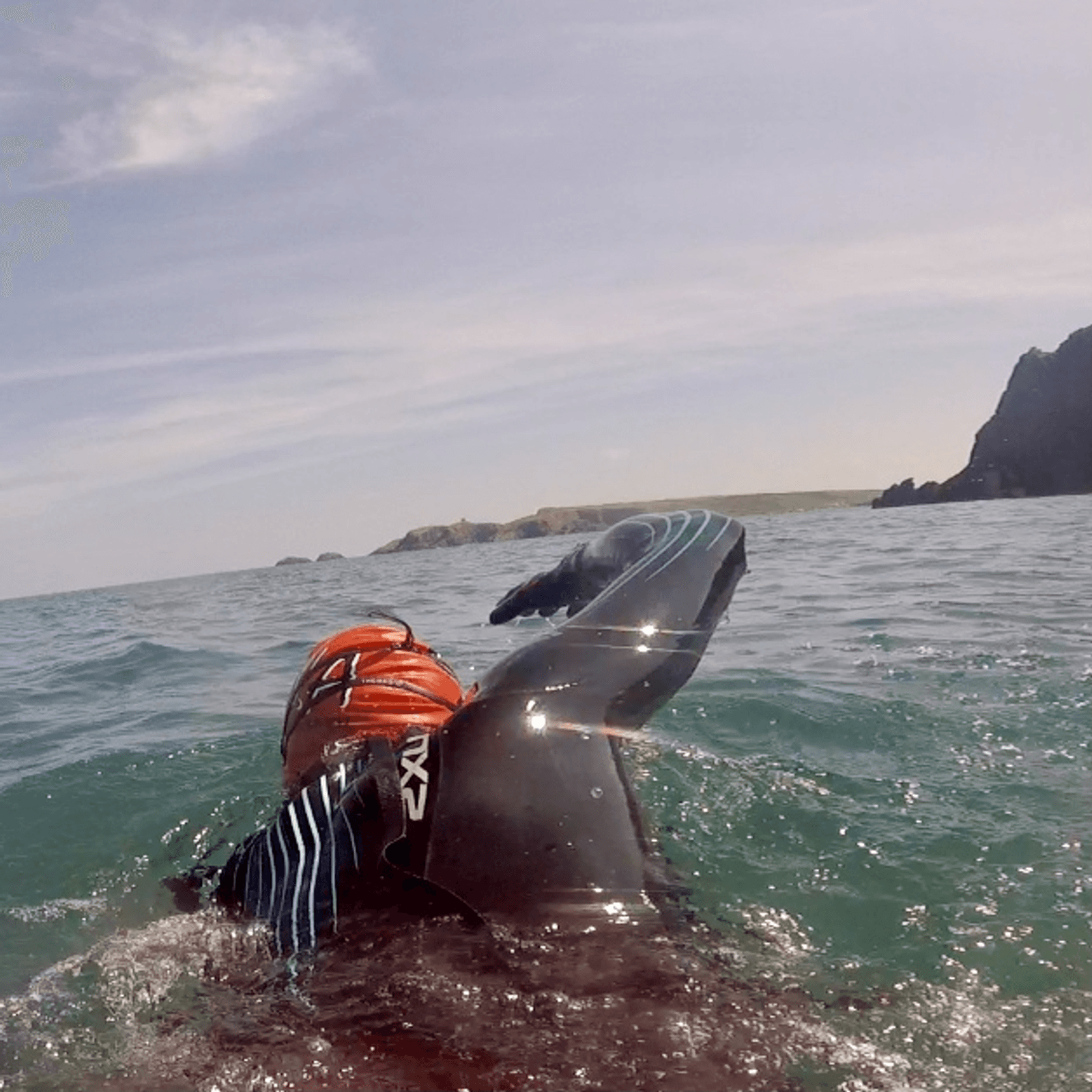 A swimmer wearing a black wetsuit and an orange swimming cap is captured mid-stroke in open water, with rocky cliffs and partly cloudy sky in the background.