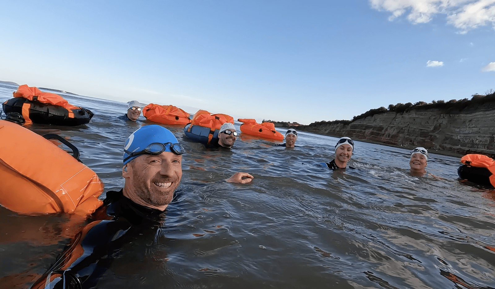 A group of people in wetsuits and swim caps, some wearing goggles, are floating in open water with orange safety buoys. They appear to be enjoying themselves, smiling and looking towards the camera. The shoreline with cliffs and partly cloudy sky is visible in the background.