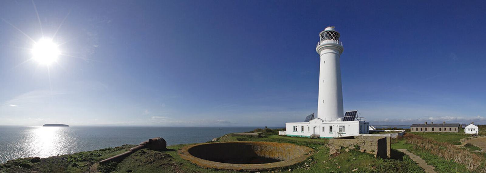 A tall white lighthouse with an adjacent building stands on a grassy cliff overlooking a calm sea under a clear blue sky. The sun shines brightly above the horizon, casting shadows. A circular concrete structure is in the foreground. Several smaller buildings are nearby.