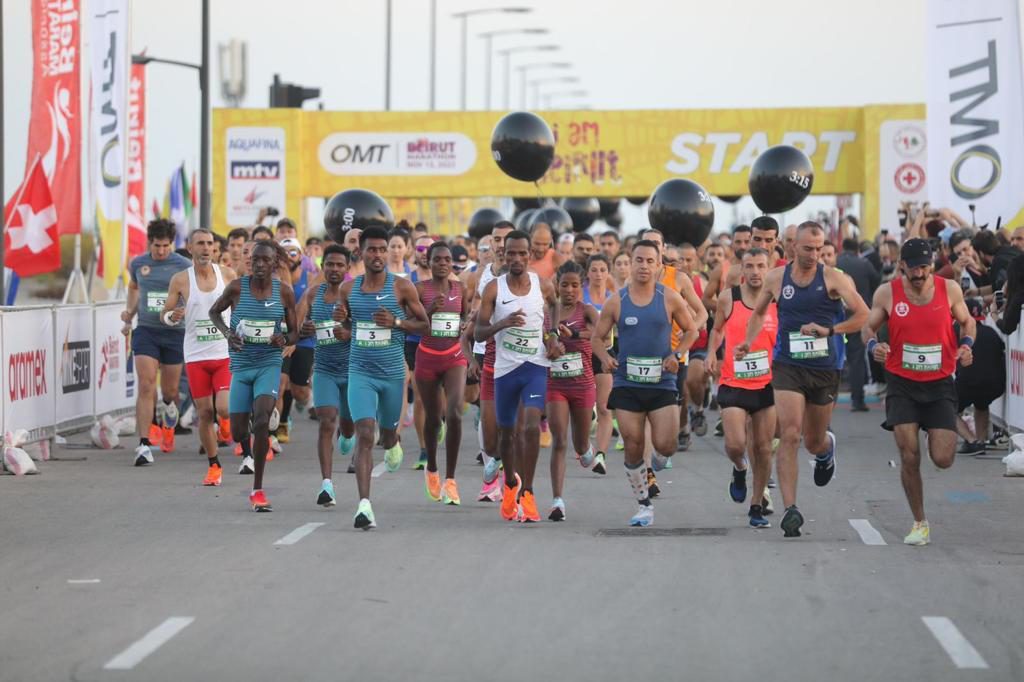 A large group of runners in athletic gear are starting a race on a road, passing under a yellow "START" banner, with race officials and spectators watching from the sides.