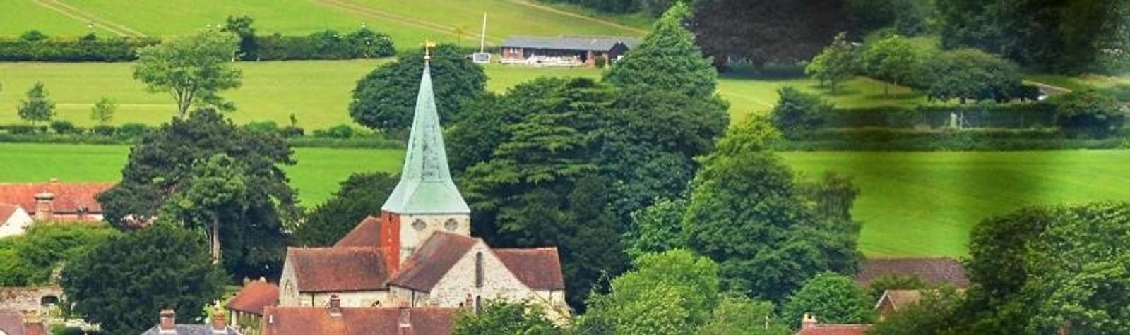 A quaint village featuring a church with a tall pointed spire surrounded by lush greenery and trees. The scene is set against a backdrop of fields and a few scattered houses, creating a picturesque, serene countryside landscape.