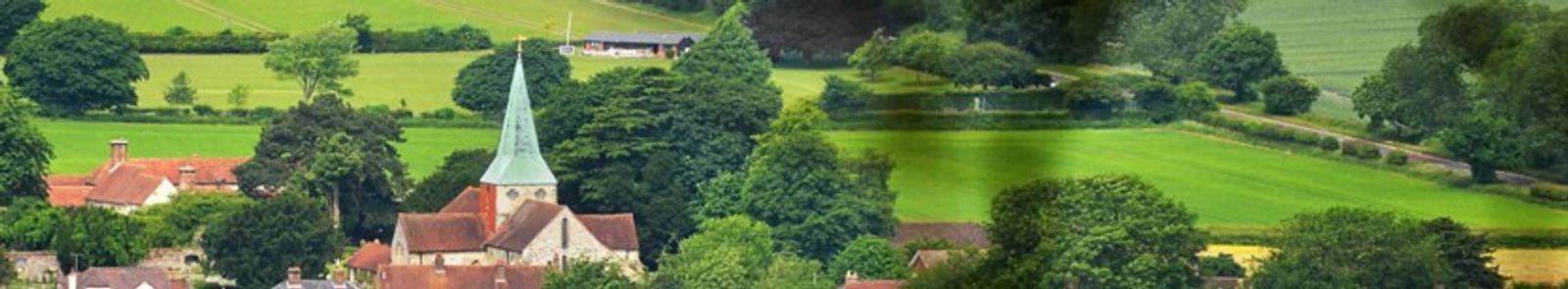 Panoramic view of a tranquil countryside village featuring a prominent church with a pointed steeple surrounded by lush green fields, trees, and scattered houses. The landscape is vibrant and serene, suggesting a peaceful rural setting.
