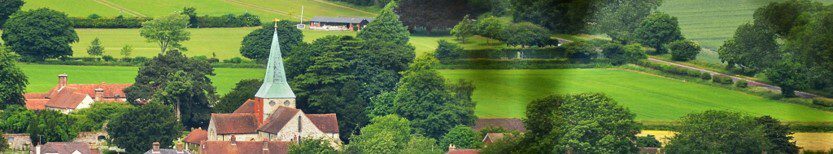Panoramic view of a tranquil countryside village featuring a prominent church with a pointed steeple surrounded by lush green fields, trees, and scattered houses. The landscape is vibrant and serene, suggesting a peaceful rural setting.