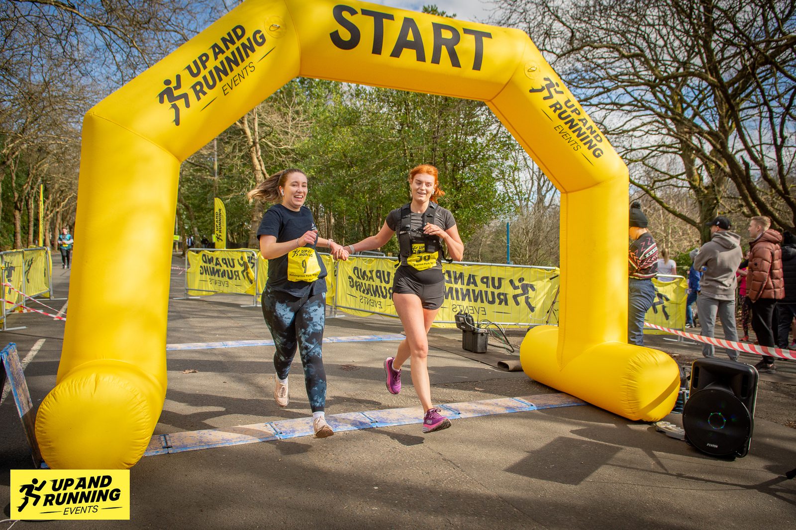 Two women are running together under a yellow inflatable "START" arch at a race event organized by UP AND RUNNING EVENTS. They are smiling and appear to be enjoying themselves. Spectators and trees are in the background.
