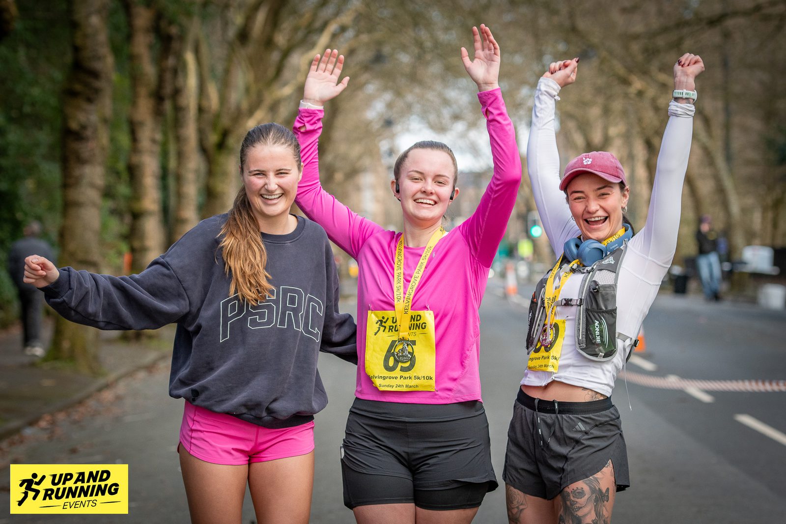 Three joyful runners with medals pose with their arms raised, celebrating their achievement. They wear athletic gear and big smiles. Behind them is a tree-lined street. A yellow banner in the corner reads "Up and Running Events.