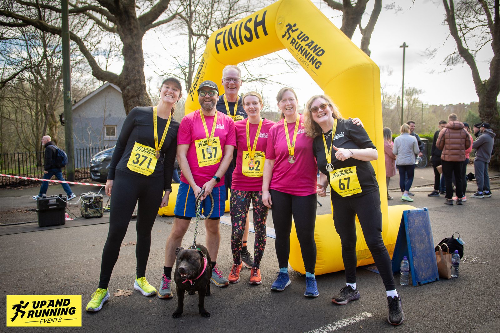 Six smiling runners, some with medals, pose under a yellow "FINISH" arch after a race. They wear bib numbers 371, 157, 82, 52, 67. A small black dog is at the front left. Trees and spectators are visible in the background. An "UP AND RUNNING EVENTS" banner is on the bottom left.