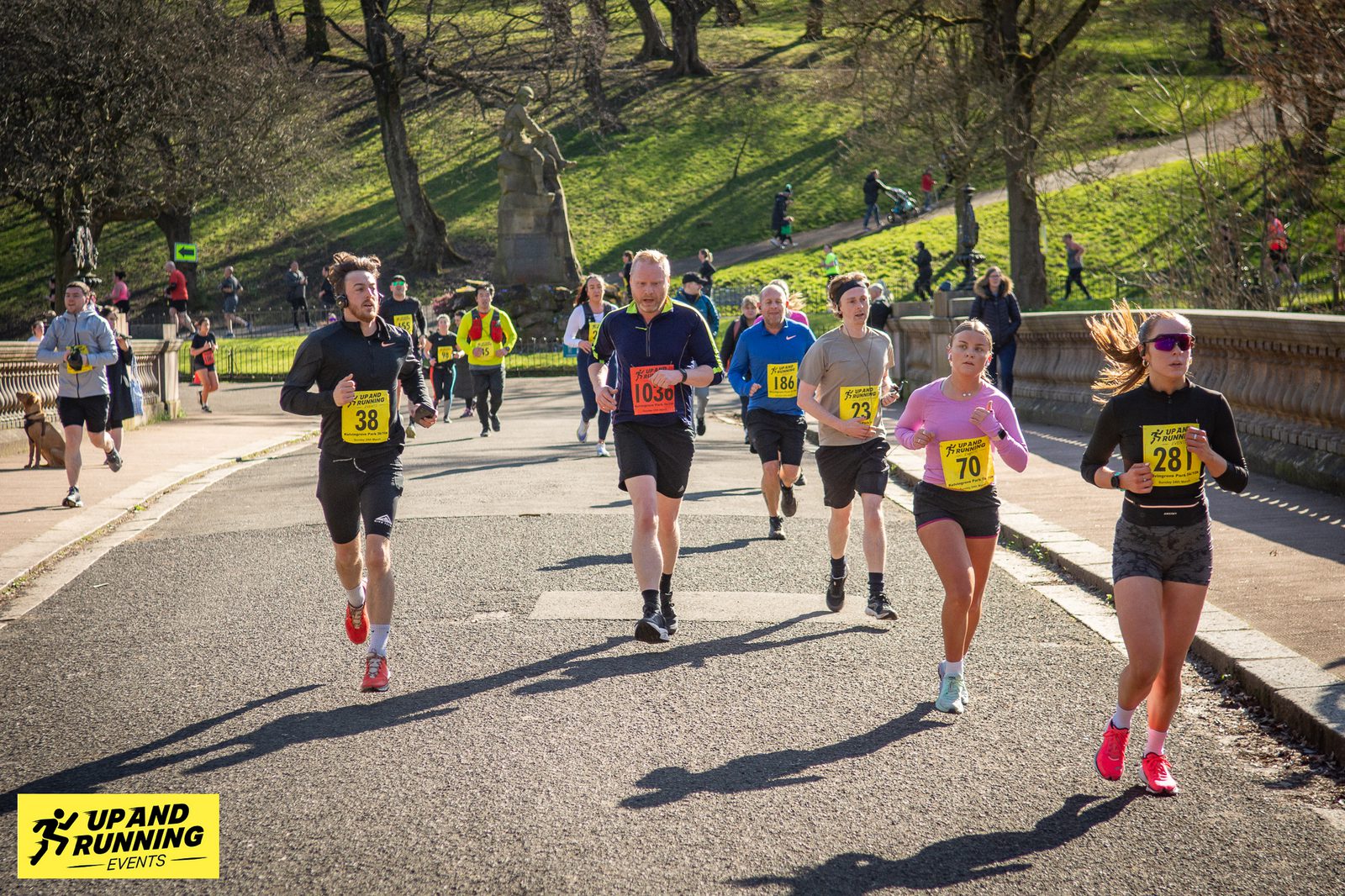 A group of runners, wearing numbered bibs, participating in a race on a paved path in a park. The sun is shining, and there are trees and a statue in the background. A sign in the bottom left corner reads "Up and Running Events.