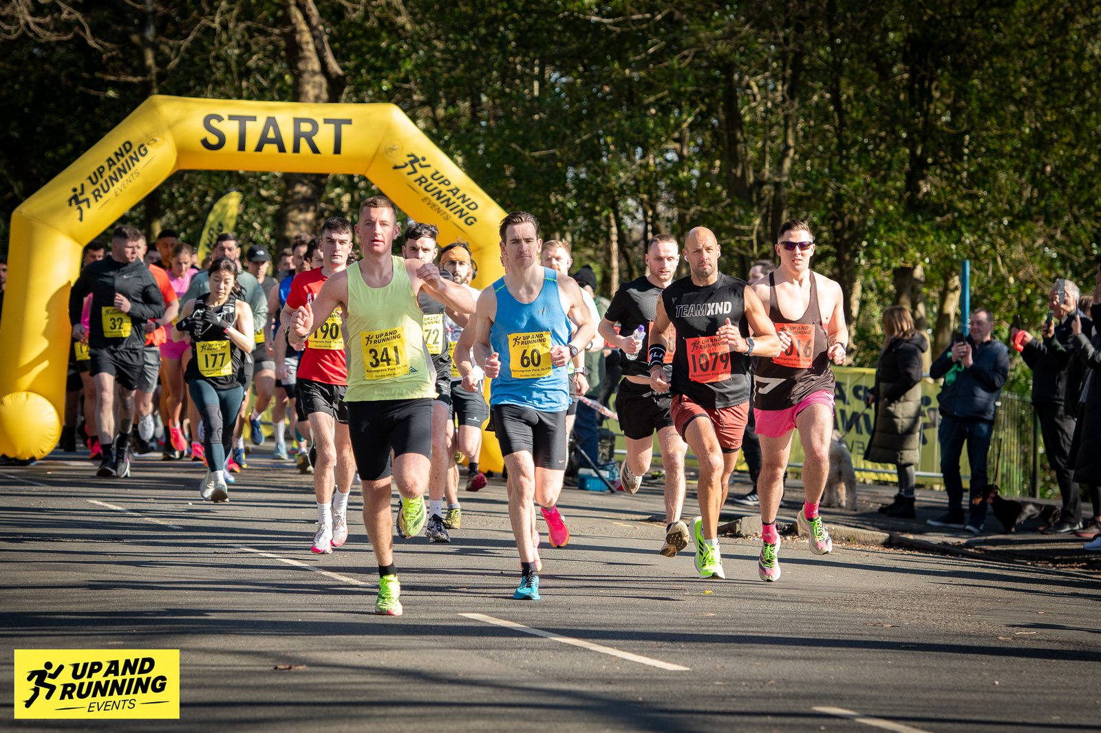 A group of runners is starting a race, passing under a yellow inflatable arch labeled "START." They are on a tree-lined street, with some wearing race bibs showing numbers. The event banner in the foreground reads "UP & RUNNING EVENTS.