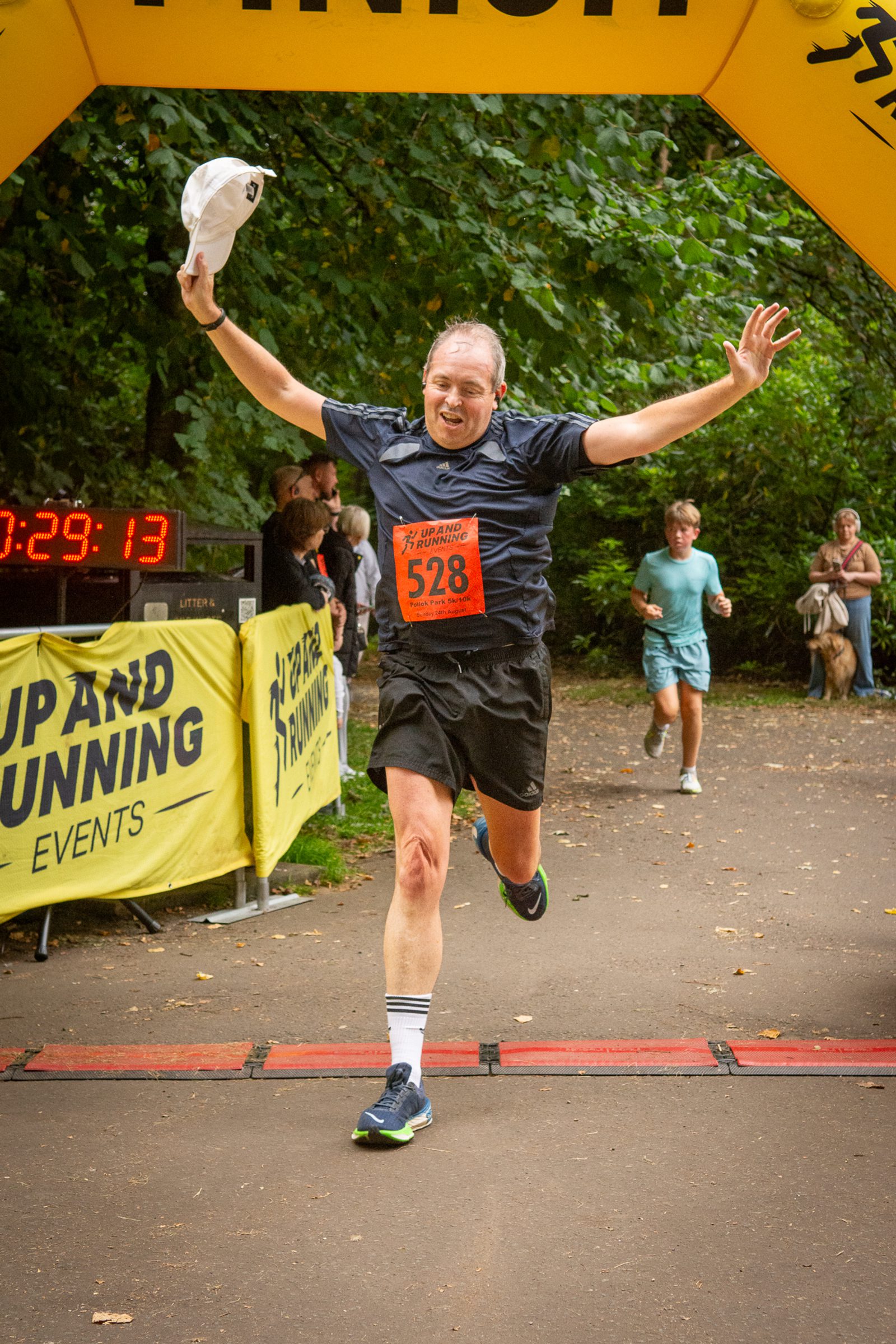 A runner in a black shirt and shorts joyfully crosses the finish line with arms raised and a cap in hand, wearing bib number 528. A digital timer and spectators are visible in the background.