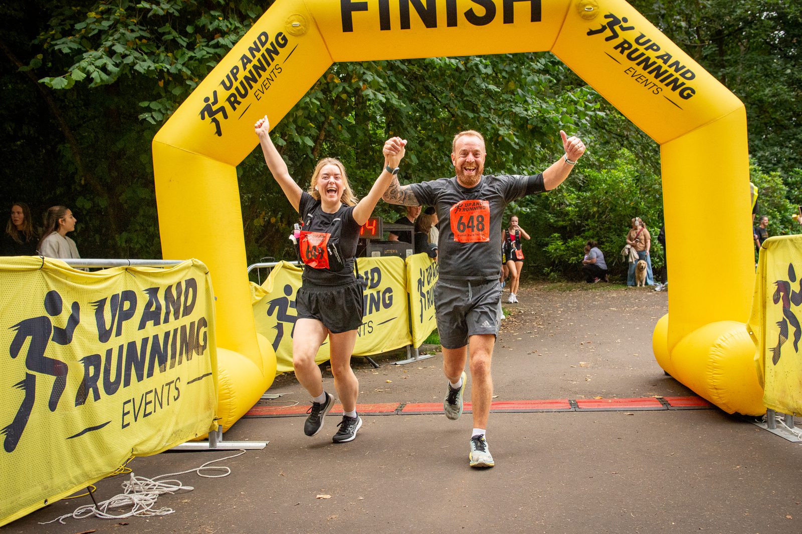 Two joyful runners, a woman and a man, cross a yellow race finish line with arms raised in celebration, wearing race bibs 646 and 648, surrounded by trees and “Up and Running Events” banners.