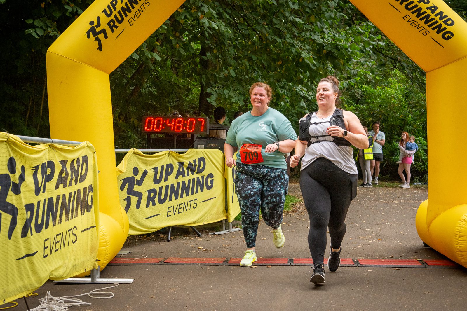 Two women smiling as they cross the finish line at a running event under a yellow "Up and Running Events" archway. A timer in the background shows 00:48:07. Spectators stand along the path.