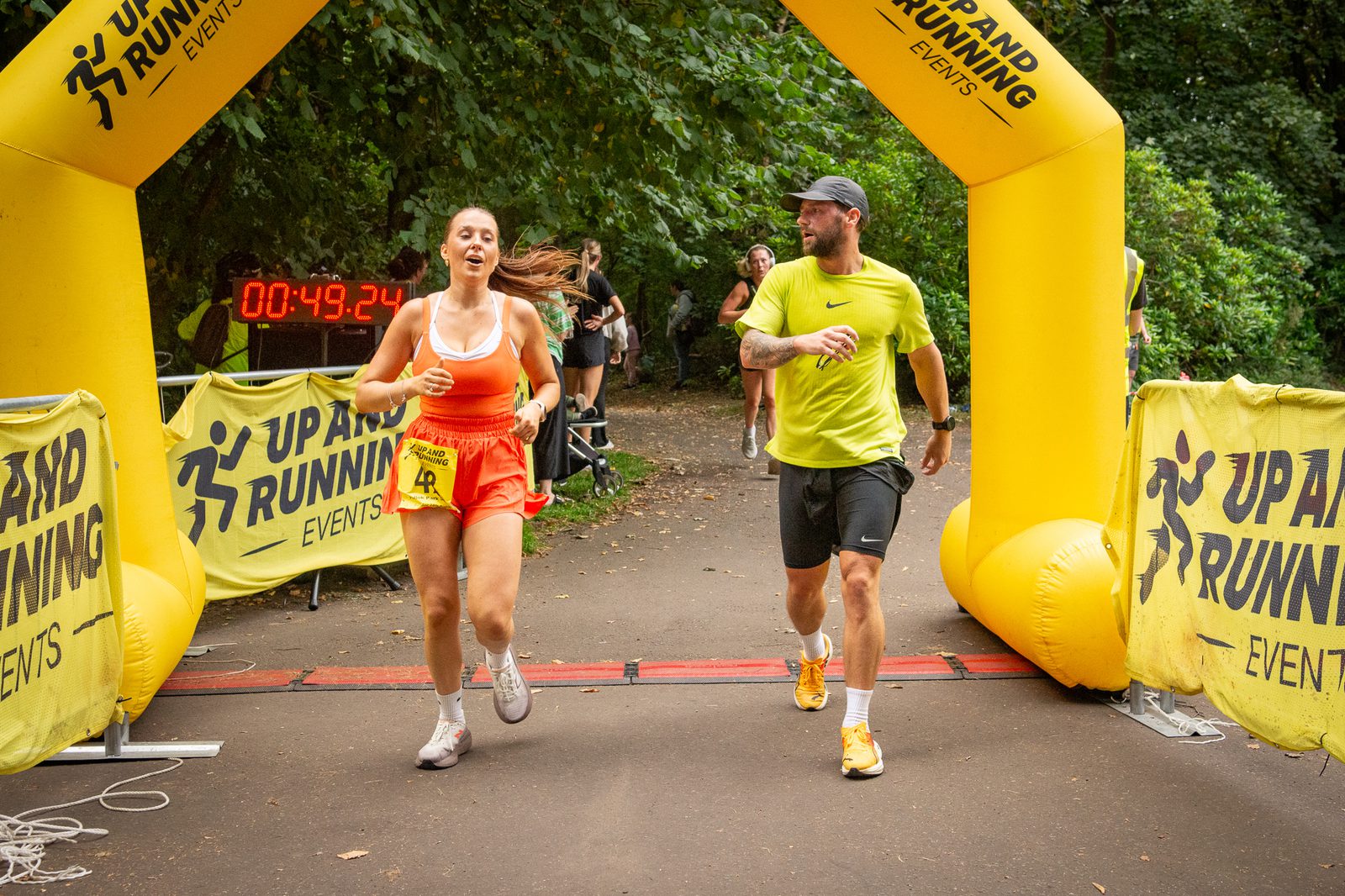 Two runners, a woman in orange and a man in yellow, cross the finish line of a race together under a yellow archway with "Up and Running Events" banners, surrounded by greenery. A timer reads 00:49:24.