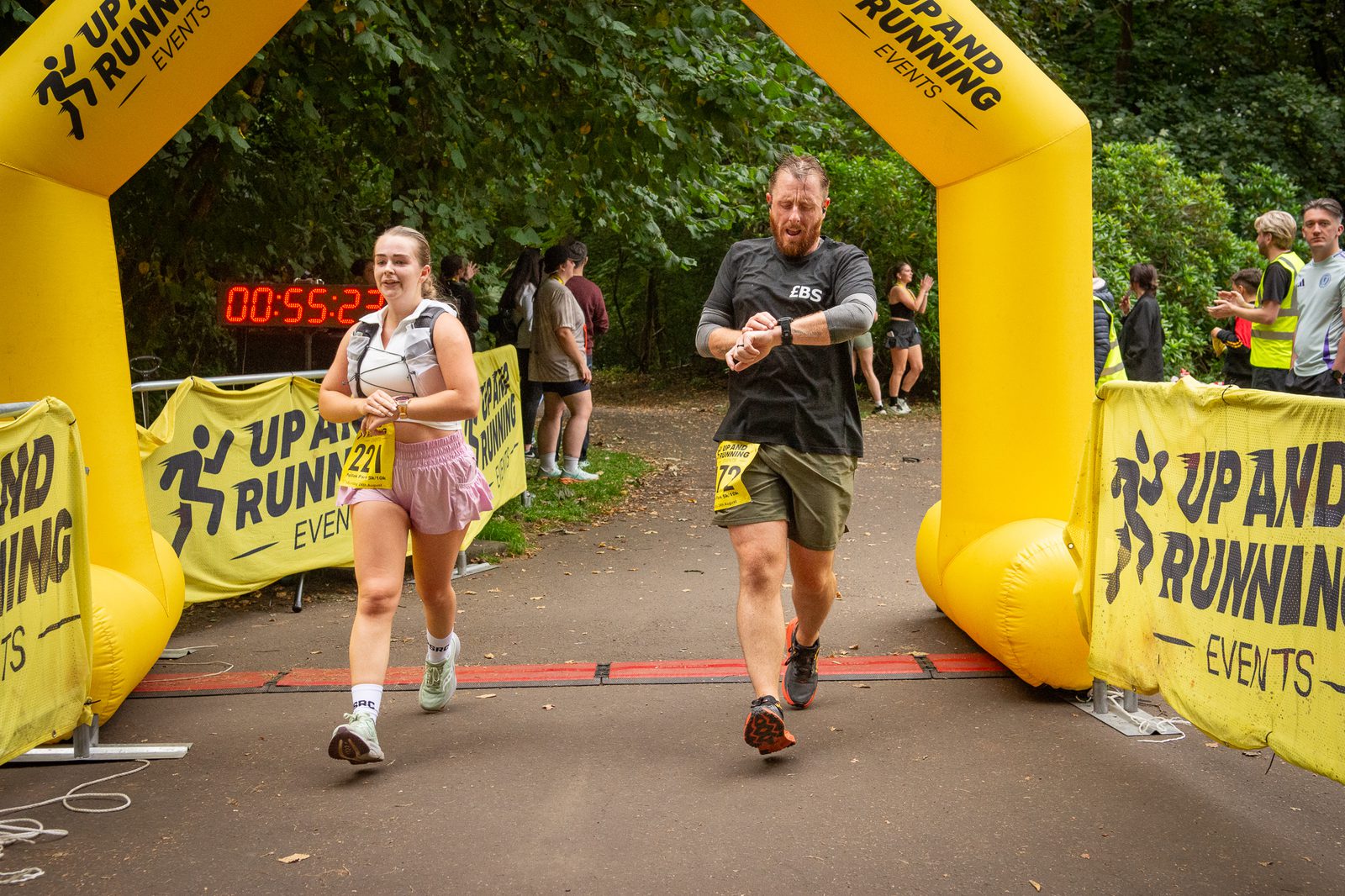 A young woman and a man cross a yellow inflatable finish line at a running event in a park. The man checks his watch, while a digital timer behind them reads 55:32. Spectators stand nearby.