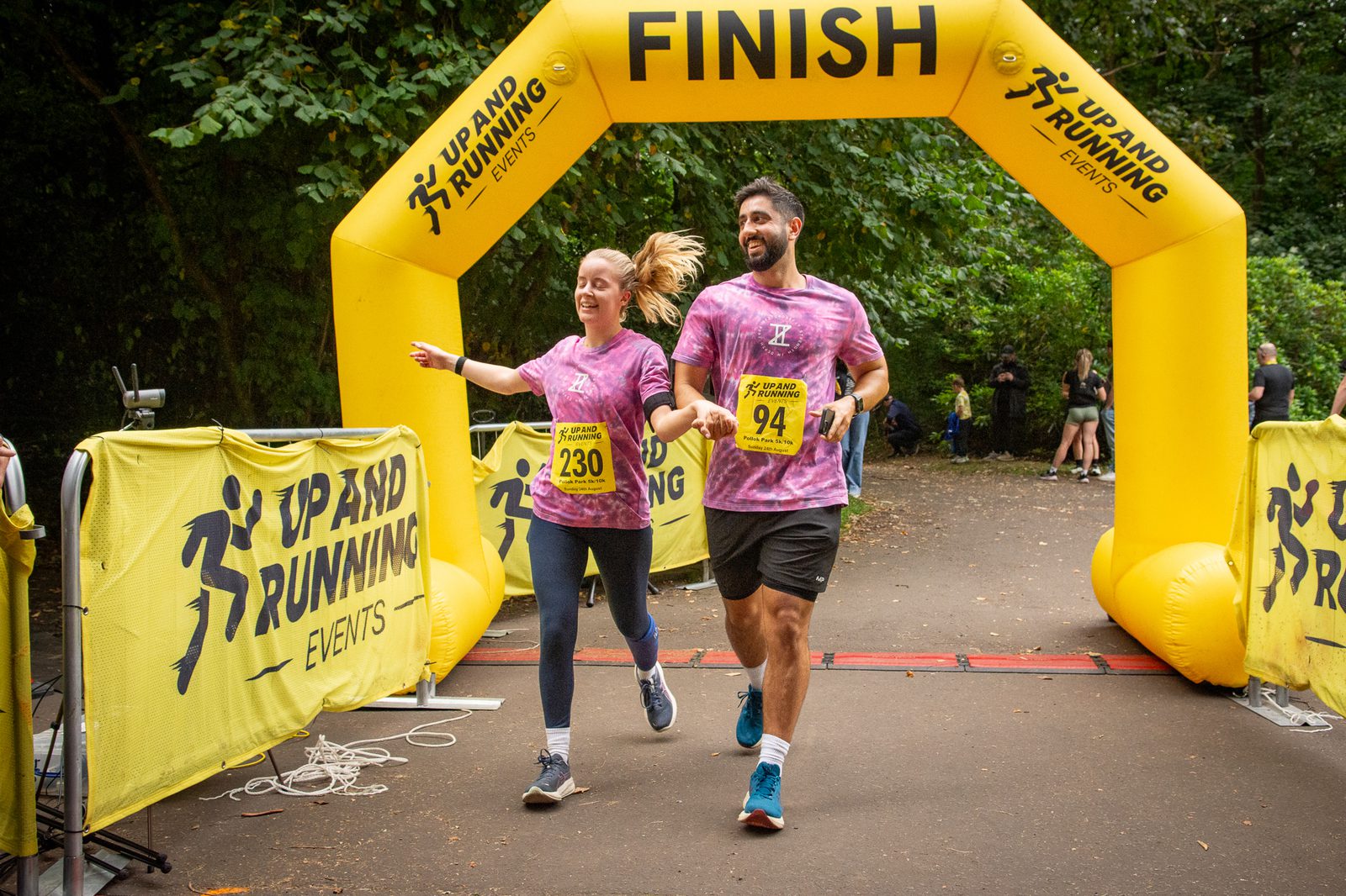 Two runners in matching purple shirts, smiling and holding hands, cross the finish line under a yellow "Up and Running Events" arch in an outdoor race. Both wear numbered race bibs and running shoes.