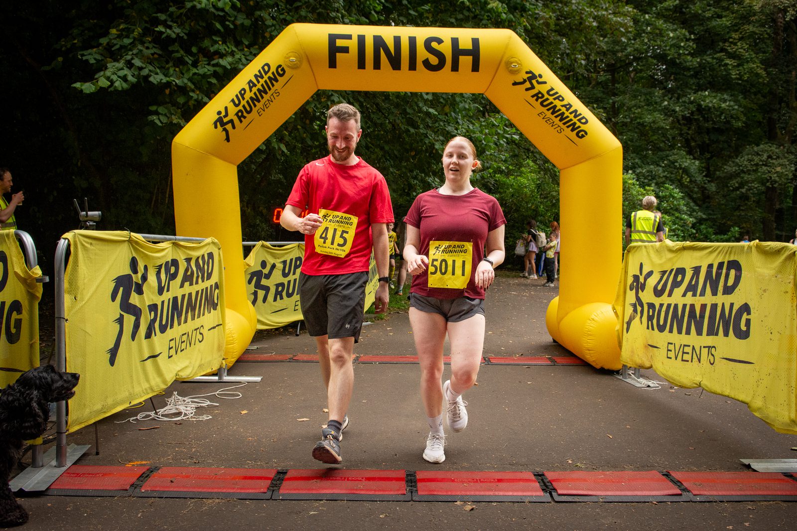 Two runners, wearing numbered race bibs, smile as they cross the finish line under a yellow "Finish" arch at an outdoor running event, surrounded by trees and banners that read "Up and Running Events.