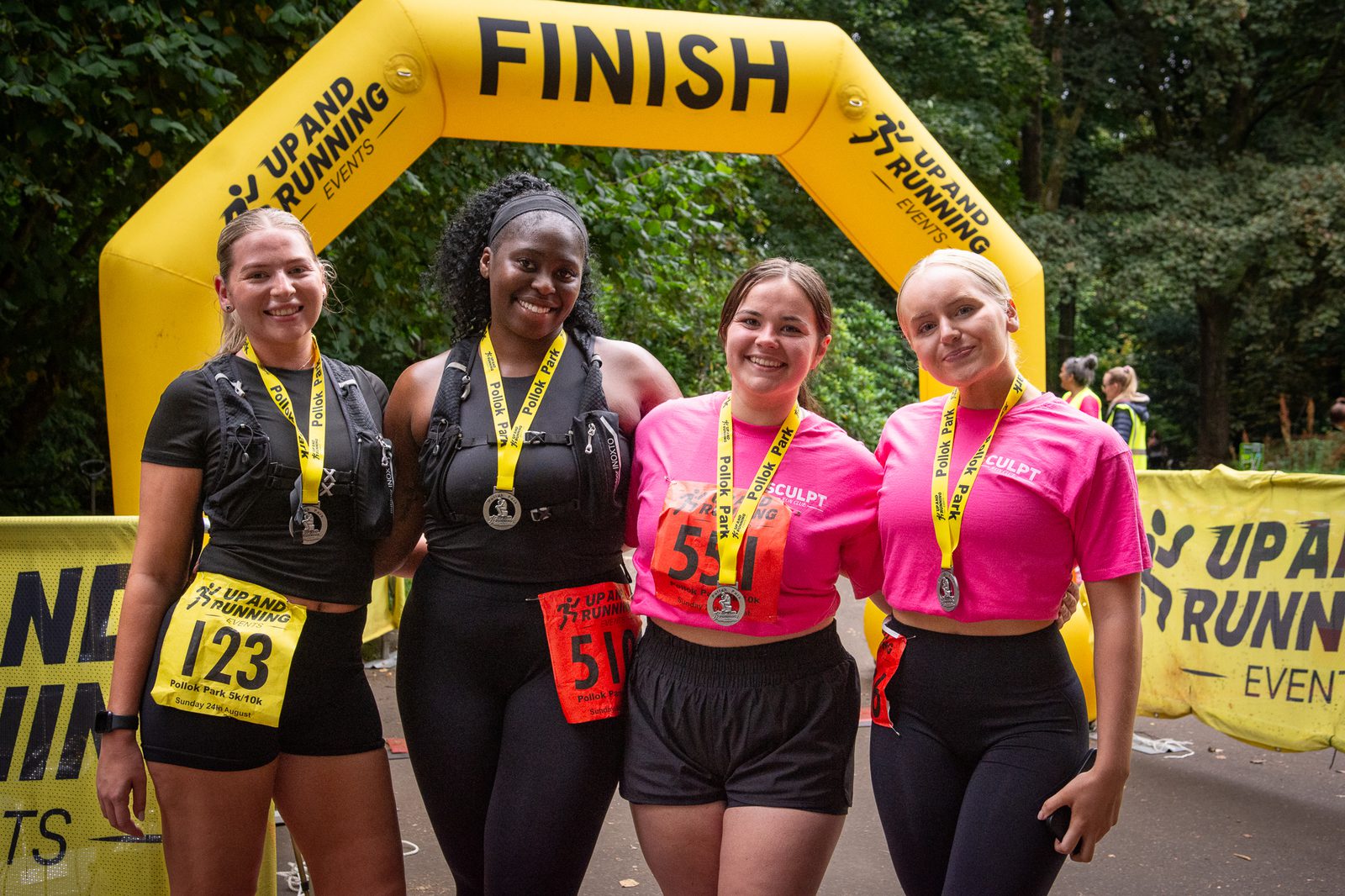 Four smiling women with medals stand under a yellow “FINISH” arch after a race, wearing running gear and numbered bibs, with trees and event banners in the background.