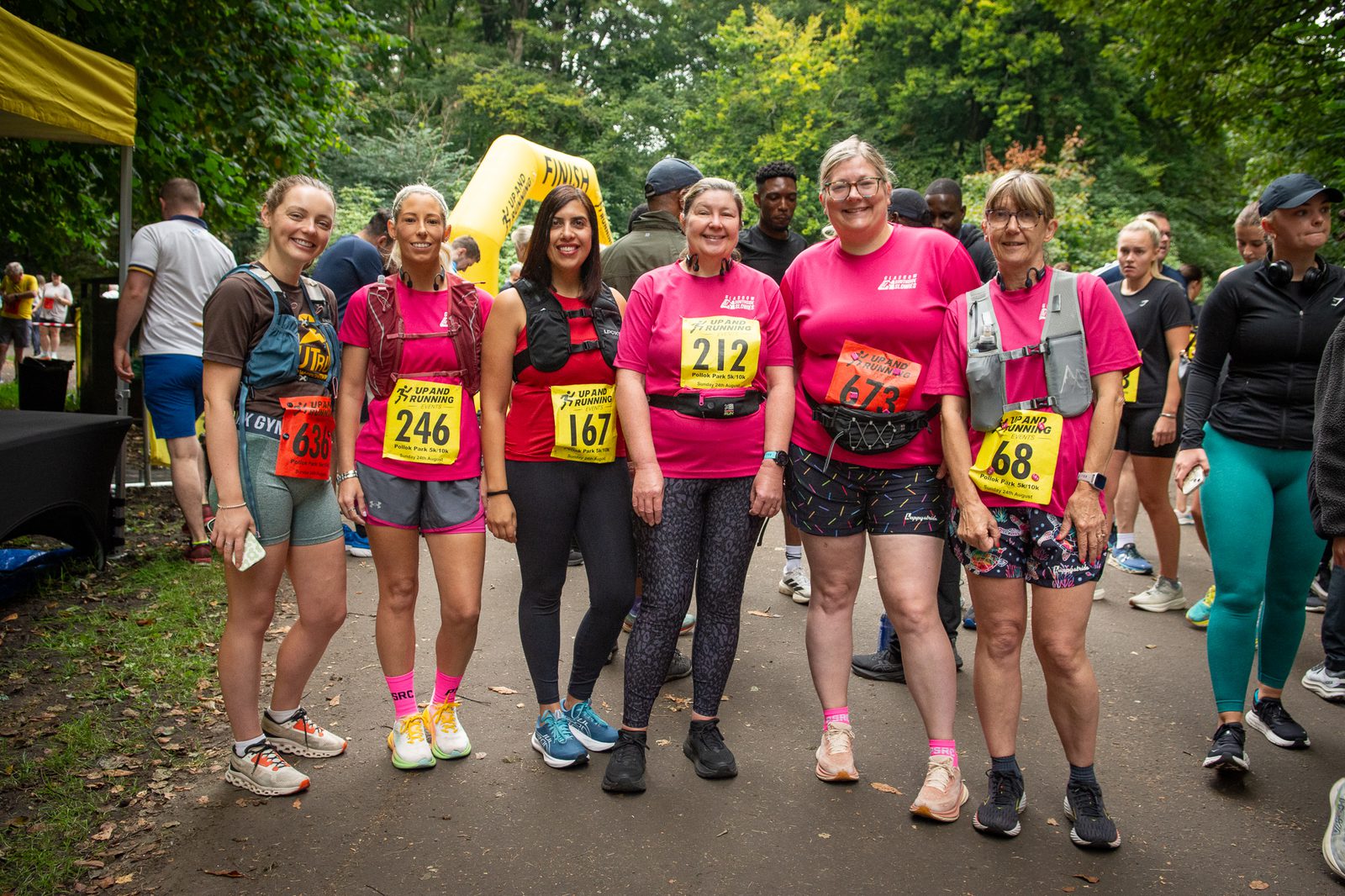 Six women in running gear, all wearing matching pink shirts and race bibs, stand together smiling outdoors at a race event. Other participants and trees are visible in the background.