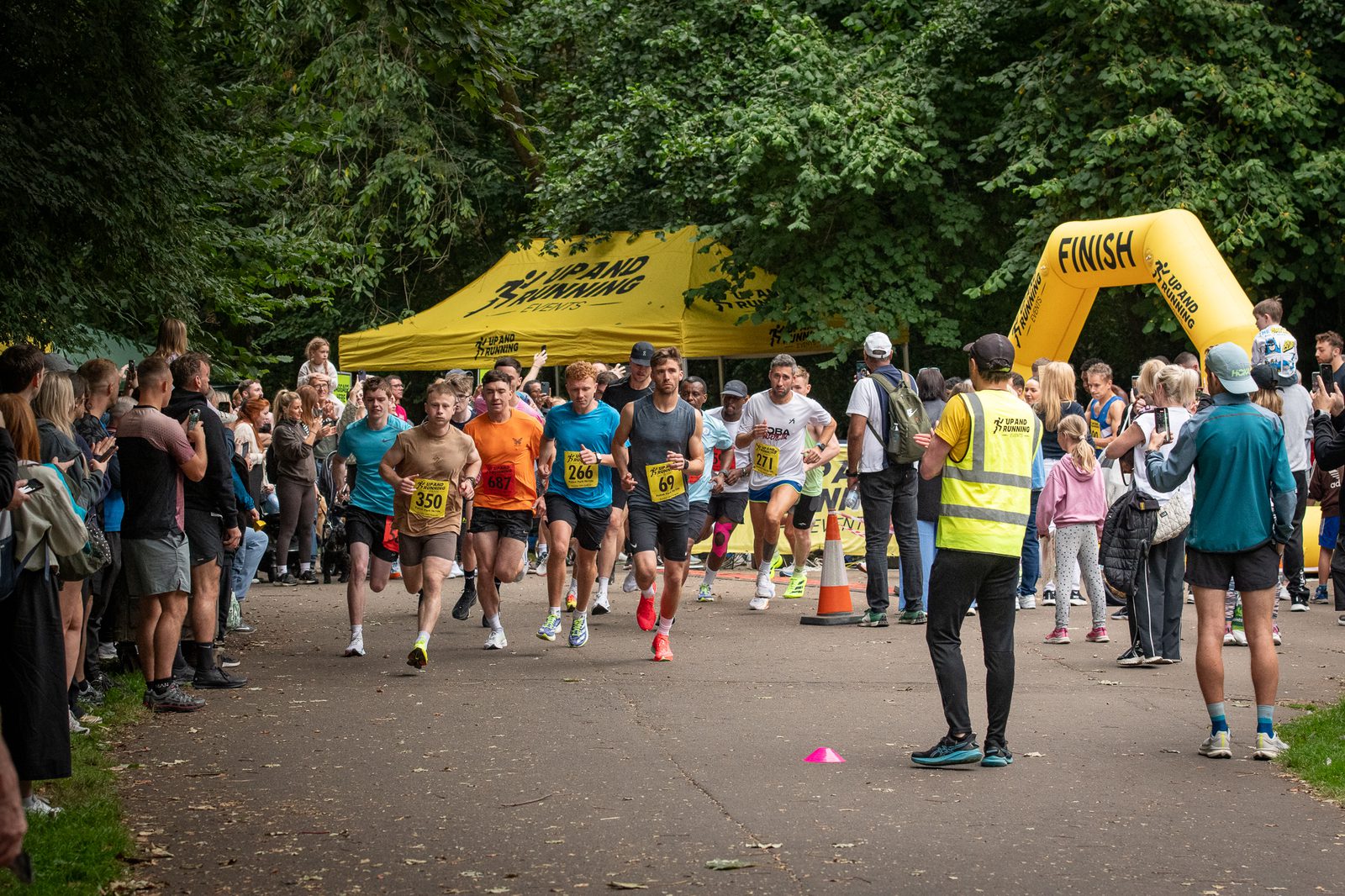 A group of runners starts a race in a park, surrounded by spectators. A yellow tent and finish line arch are visible, with a volunteer in a yellow vest standing near the runners. Trees line the path.
