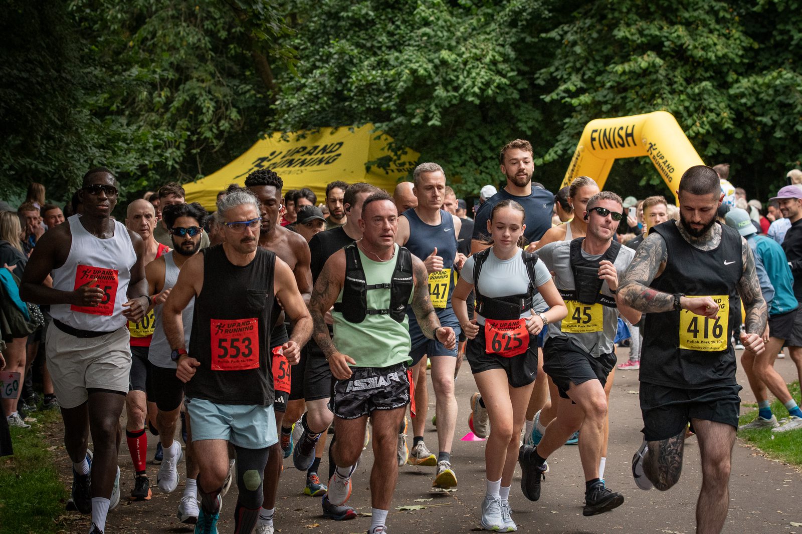 A group of runners wearing numbered bibs participate in a race on a park path, with trees in the background and a yellow "FINISH" inflatable arch visible behind them.